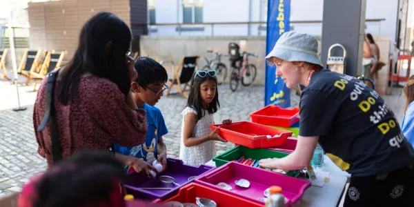 A family group of 3 with a We The Curious staff member, looking at things in a series of coloured trays in an outdoor space