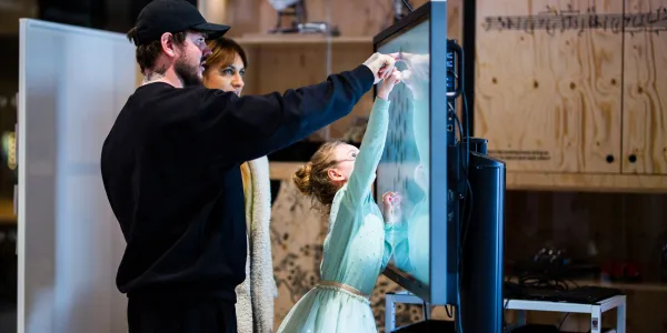 Two adults are helping a child who is reaching up and pointing at something on a screen.