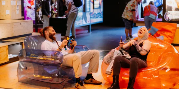 Two people are sat in the theatre of curiosity on bubble chairs, playing bopit and laughing.