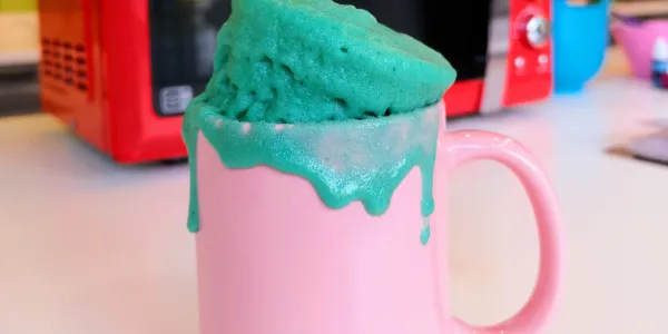 A pink mug is in front of a microwave with a bright blue mug cake spilling out of it onto the sides.