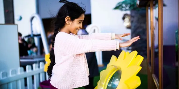 A child is stood in We The Curious science centre in a pink jumper. Stood Infront of a large, yellow, plastic flower and they are holding their hands up over it as it blows wind in their direction.