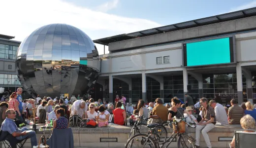 A crowd of people sat in Millennium square looking at a big screen on the We The Curious building