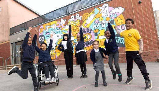 A cheerful group of children in school uniform in front of a bright colourful mural on a brick wall. They're all smiling, with arms in the air and some mid-jump