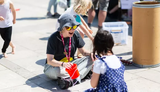 A Live Science Team member sat down outside in the sun, cross legged towards the camera. She has a small red bucket in her hand and is wearing sunglasses and a sun hat. She is smiling enthusiastically opposite a young girl who is wearing a blue polka dot dress facing away from the camera.