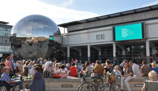 An outside space with a large electronic screen on a building, with hundreds of people sat watching 