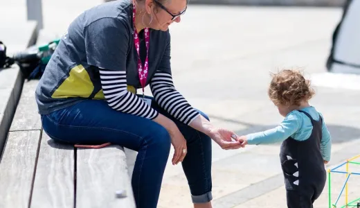 An adult and toddler playing with colourful straws together. The toddler is putting something in the adult's hand, they're outside in a stone paved space, with the adult sat on a wooden bench. 