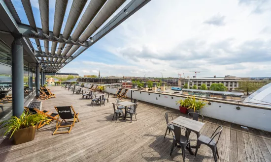 A large terrace with wooden decking overlooking Millennium Square. There are tables, chairs and plants placed at equal distance throughout.