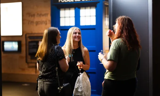 A group of adults with champagne in hand standing and talking in the We The Curious venue. The blue Tardis can be seen in the background. 