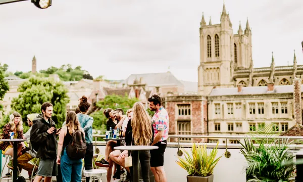 A group of young adults gathered around on the terrace, smiling and talking. A view of The Bristol Cathedral is in the background. 