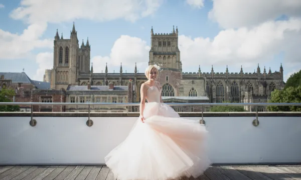 A woman in a flowing wedding dress standing on a large balcony posing in front of Bristol Cathedral 