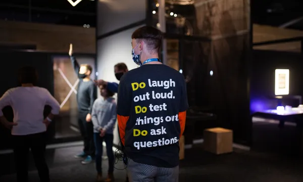 A We The Curious Live Science Team member standing in the venue, facing away from the camera with the words, 'Do laugh out loud. Do test things out. Do ask questions.' Printed on the back of their uniform