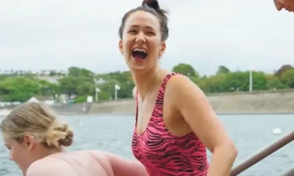 A woman in a swimming costume is turning to the camera and laughing. There is a body of water behind her and some trees in the distance. Another woman is in front of her but not fully in view.