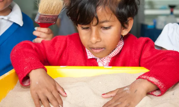 A child with their hands in a tub of sand, a hand holding a brush is by their right shoulder