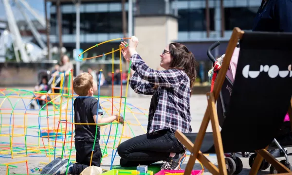 Grown-up and a child building a colourful structure out of plastic straws on the sunny Millennium Square 