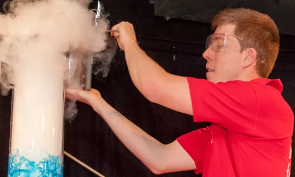 a man in a red top next to a tube with blue liquid, white vapour is billowing out the top of the tube