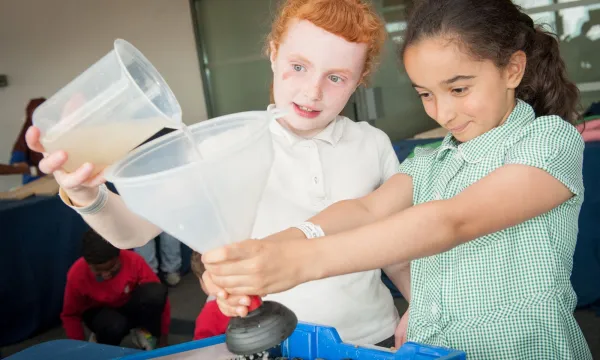 a student holds a funnel in outstretched arms while another student pours water in from a jug