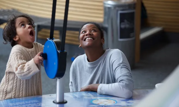 A woman looks up smiling. She is next to a young child with a look of amazement on their face. They are playing with a blue exhibit in We The Curious