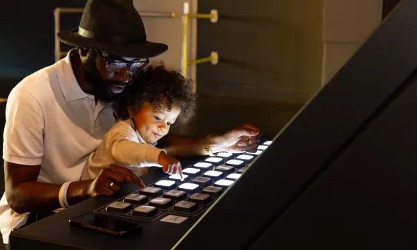 A child is sat on an adults lap and they are pressing lit up buttons on an exhibit in a science centre.