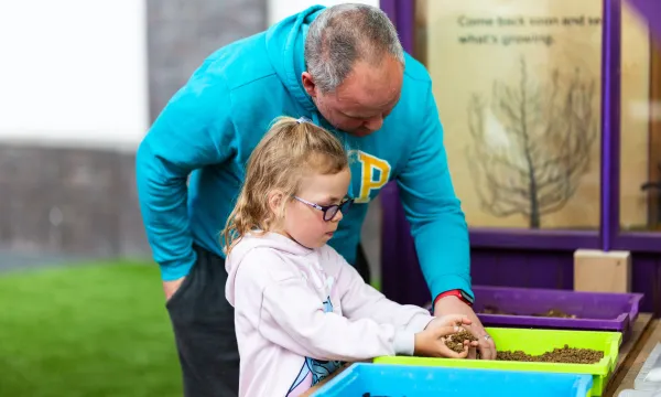 An adult and a child are in a greenhouse playing with soil in plastic containers.