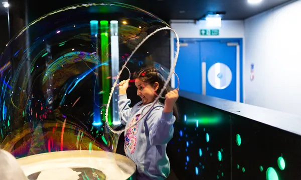 A child is in the middle of a science centre in a jumper pulling a sparkling bubble over their head.