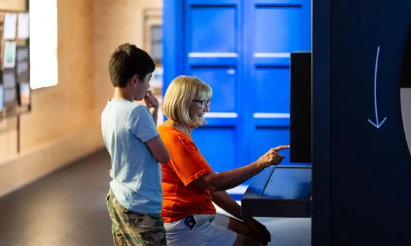 An adult and a child are in a science centre. The adult is sat down pointing at a screen and the child is stood next to them watching.