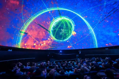 A shot of people sat in the Planetarium, a cinema-like experience. On the domed-shaped screen there's a visual of a colourful nebula.