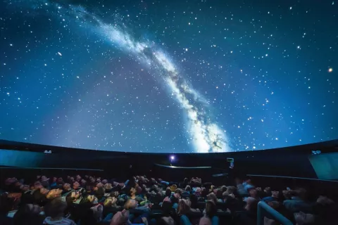 Audience in a planetarium watching a projection of the Milky Way galaxy on the dome-shaped ceiling.