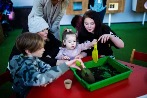 A family with a young child at the centre, sat around a red table with a tray of soil in the middle.