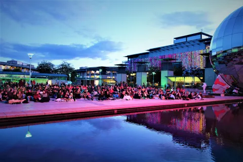 A big ground sitting in deckchairs and on the floor on a public square enjoying a screening of Romeo and Julia on a big screen outside We The Curious