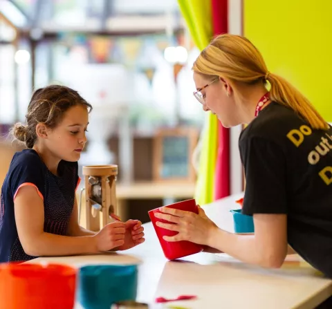 An adult and child are together in a kitchen space with brightly coloured bowls on a white surface. The adult is on the right, holding a red pot in their hands, which both the adult and child are looking into. The adult is wearing a grey ‘We The Curious’ uniform t-shirt and pink lanyard.