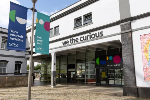 A view of the We The Curious building showing the main doors with the name and logo above. There is a colourful flag in the foreground that reads 'see your soul'.