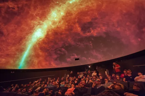 People sat in a planetarium looking up at a red sky projected on the dome.