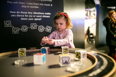 A child engages with an interactive display at a science museum, featuring colorful blocks with symbols, placed on a round table. A sign encourages visitors to explore how vibrations translate into sound.