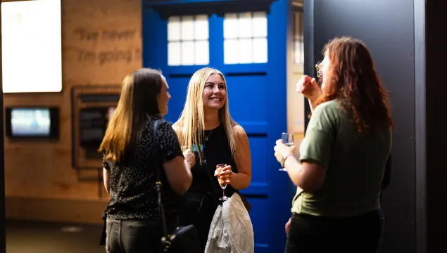 A group of three women standing amoung the exhibits talking and laughing with drinks in-hand