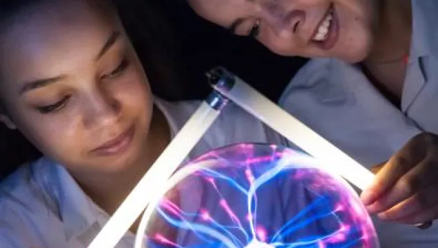 Two children each holding a small light tube over a plasma ball which is causing the tubes to emit light.