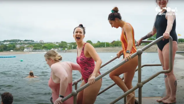 Three women in bathing costumes walking down steps into a body of water. The girl in the middle has her head turned towards the camera and is laughing.