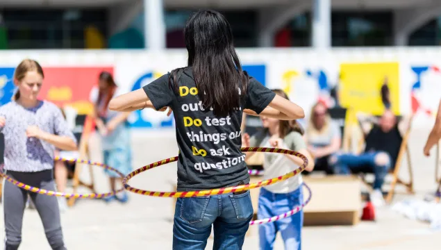A We The Curious staff member facing away from the camera, hulahooping. Their t-shirt reads 'Do test things out, Do ask questions' 