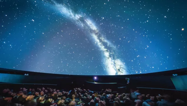 Audience in a planetarium watching a projection of the Milky Way galaxy on the dome-shaped ceiling.