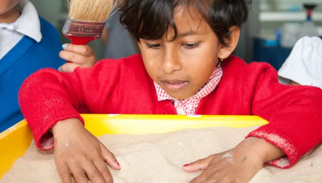 A child with their hands in a tub of sand, a hand holding a brush is by their right shoulder