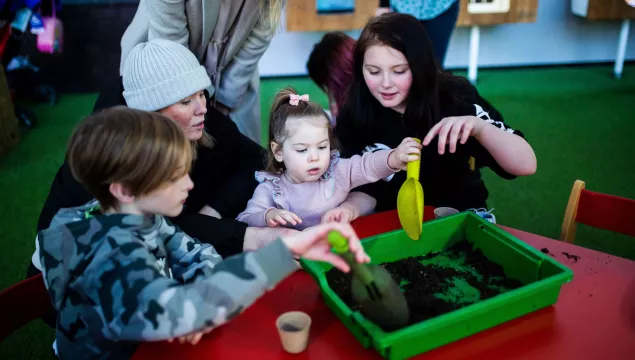 A family with a young child at the centre, sat around a red table with a tray of soil in the middle.