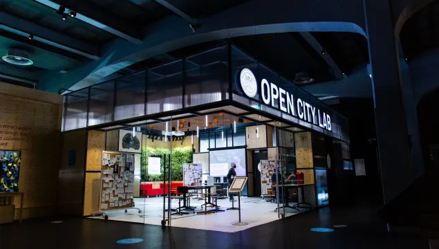 An brightly lit open space with tables, boards and activities with the name 'Open City Lab' space illuminated above 