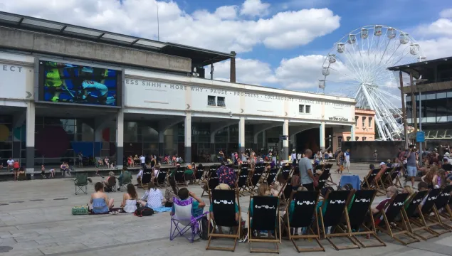 A big ground sitting in deckchairs and on the floor on a public square enjoying a screening of hockey on a big screen outside We The Curious