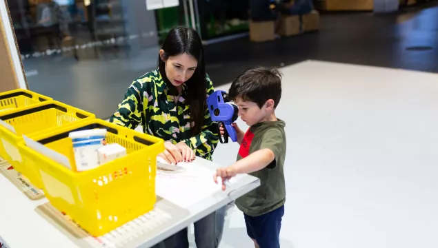 An adult and young child play with a hand-held pricing machine in the Open City Lab at We The Curious. There are yellow shopping baskets with some food containers in them on the table in front of them.