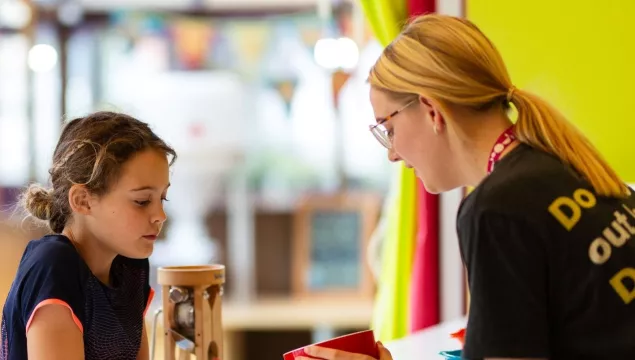 An adult and child are together in a kitchen space with brightly coloured bowls on a white surface. The adult is on the right, holding a red pot in their hands, which both the adult and child are looking into. The adult is wearing a grey ‘We The Curious’ uniform t-shirt and pink lanyard.