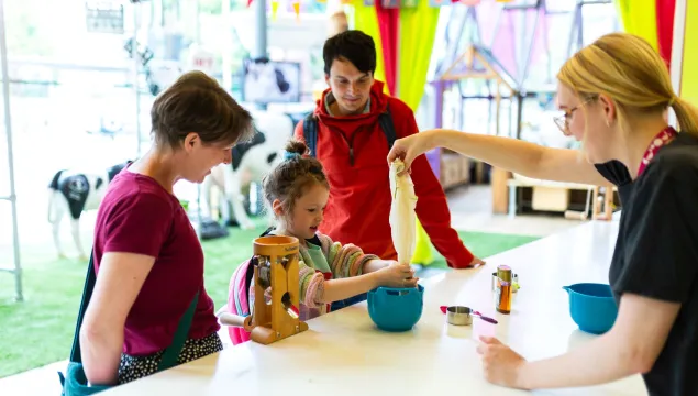 A child and two adults in the Kitchen at We The Curious. The child is being helped to make oat milk by a member of staff.