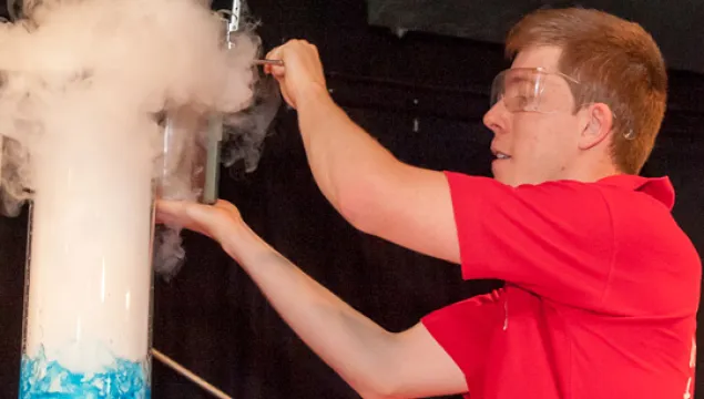 a man in a red top next to a tube with blue liquid, white vapour is billowing out the top of the tube