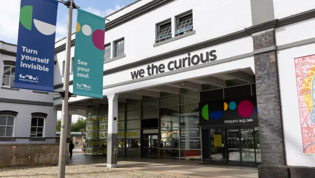 A view of the We The Curious building showing the main doors with the name and logo above. There is a colourful flag in the foreground that reads 'see your soul'.