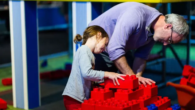 An adult and a child build a structure with large red bricks. 