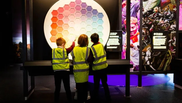 A group of children in high vis jackets look at the colour wheel at We The Curious, with their backs to the camera.