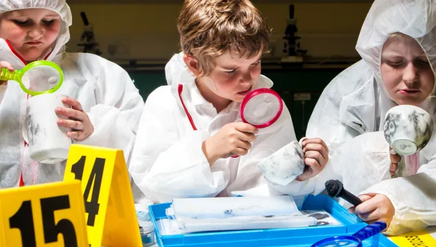 Students in white overalls look at fingerprint covered mugs with magnifying glasses